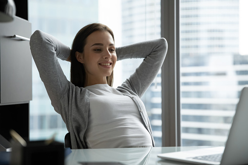 Smiling young woman employee sit at desk stretch at workplace look at laptop screen relaxing, happy millennial woman worker rest in chair at office table watch webinar on computer online