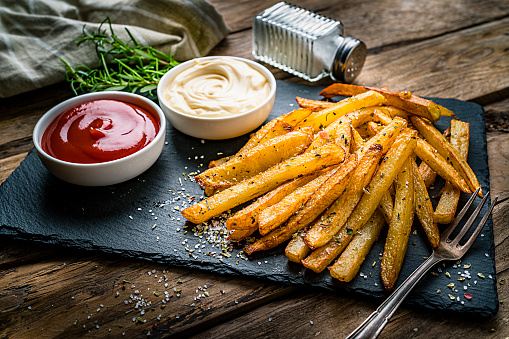 Homemade french fries served with ketchup and mayonnaise placed on a slate board shot on rustic wooden table. Predominant colors are yellow and brown. High resolution 42Mp studio digital capture taken with SONY A7rII and Zeiss Batis 40mm F2.0 CF lens