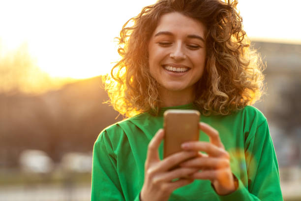 Young hipster woman using smartphone at sunset Beautiful young woman with curly hair smiling and using smartphone city of mobile stock pictures, royalty-free photos & images