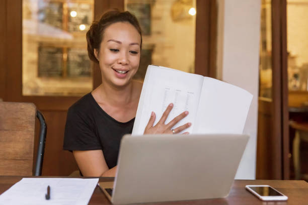 Happy Asian woman teaching online class from home during Covid 19 pandemic Portrait shot of happy online teacher holding a study book, while teaching in front of her laptop at home , during quarantine Covid 19 e learning photos stock pictures, royalty-free photos & images