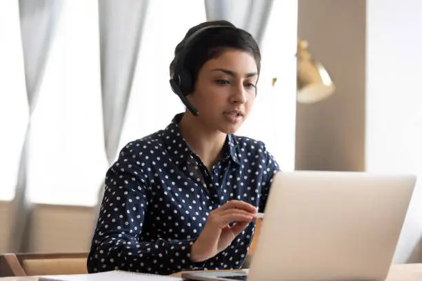 Serious Indian woman wearing headphones with microphone talking, using laptop, looking at screen, young female consulting client, video call, student learning language online, listening lecture