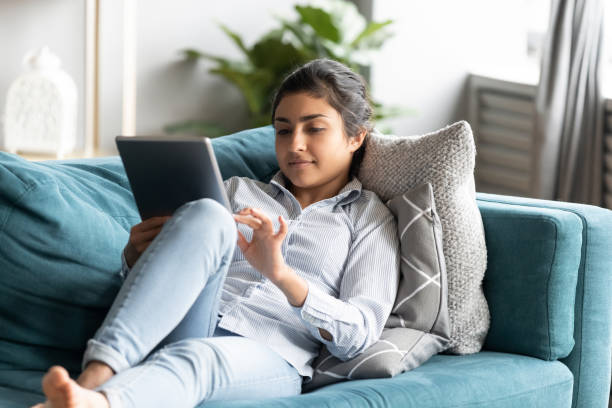 Peaceful Indian woman using computer tablet, relaxing on couch