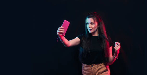 istock Young girl posing with smart phone in her hands, making selfie on black background. Studio shots, copy space for text 1218229010
