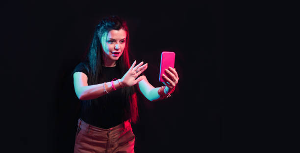 istock Young girl posing with smart phone in her hands, making selfie on black background. Studio shots, copy space for text 1218229051