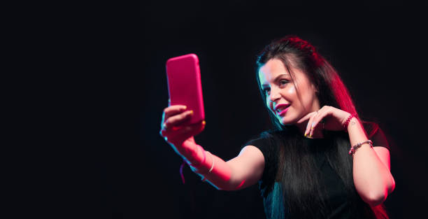 istock Young girl posing with smart phone in her hands, making selfie on black background. Studio shots, copy space for text 1218229065