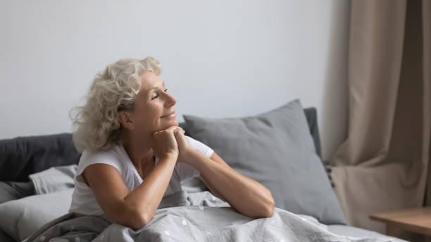 Peaceful middle aged woman welcoming new day in bedroom. Peaceful calm positive middle aged senior retired woman sitting in bed after wakeup in weekend morning, enjoying good mood after good night rest relaxation, welcoming new day alone in bedroom. tranquil evening stock pictures, royalty-free photos & images