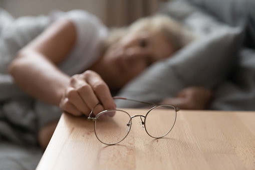 Close up focus on eyeglasses on wooden bedside table. Elderly senior retired woman suffering from blurred eyesight, taking optical eyewear after waking up in bedroom, bad vision ophthalmology concept.