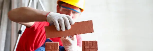 Close-up of professional builder repairing house. Professional constructor laying bricks. Man in special uniform and helmet. Restructuring and construction concept
