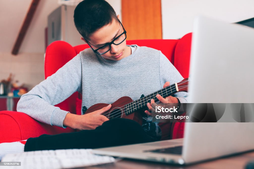 The kid in the red armchair learns to play ukuleles during isolation due to the coronavirus The kid in the red armchair learns to play ukuleles during isolation due to the coronavirus (Stay home concept) Learning Stock Photo