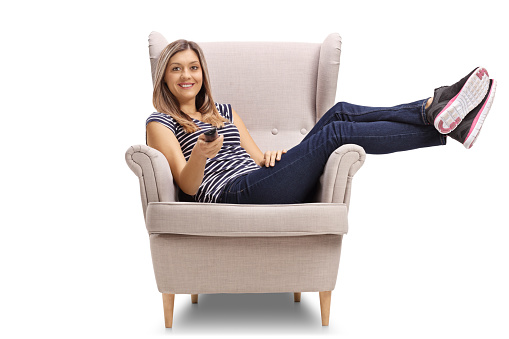 Young woman sitting in a cosy armchair and holding a remote control isolated on white background