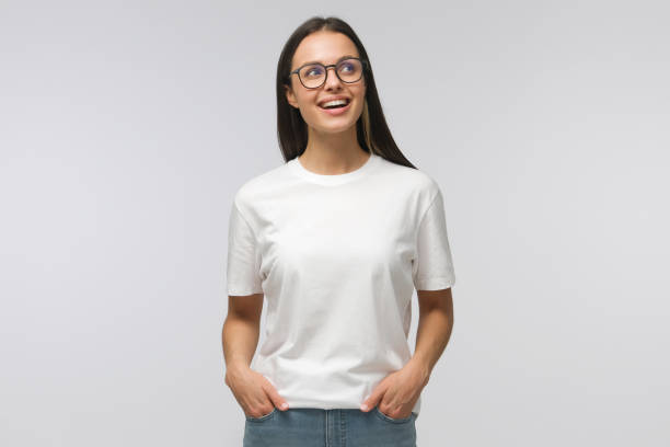 白いtシャツとジーンズに身を包み、眼鏡をかけ、ポジティブな笑顔で脇を見て、灰色の背景に孤立した幸せな学生の女の子 - shirt caucasian white one person ストックフォトと画像