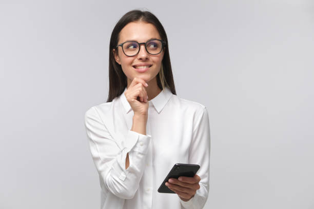 Cunning woman making plans while holding cellphone in hand and looking to right side through big glasses with smile, isolated on gray background with copy space