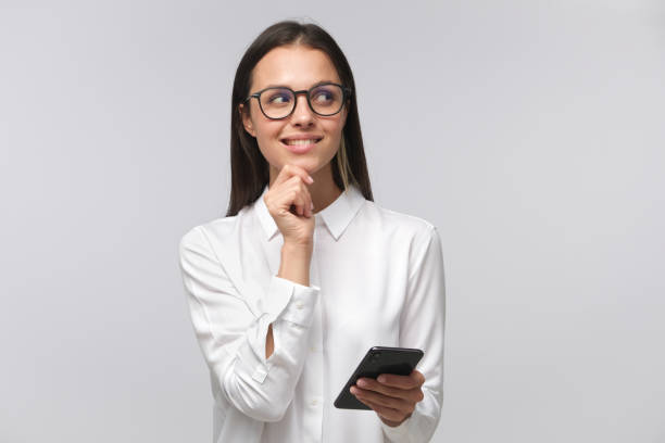 Cunning woman making plans while holding cellphone in hand and looking to right side through big glasses with smile, isolated on gray background with copy space Cunning woman making plans while holding cellphone in hand and looking to right side through big glasses with smile, isolated on gray background with copy space conspiracy photos stock pictures, royalty-free photos & images