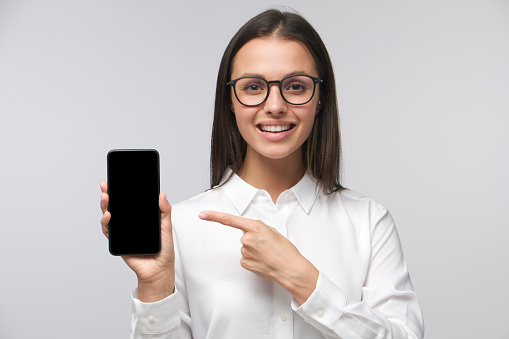 Formally dressed girl in big glasses pointing finger at blank screen of smartphone with positive smile, copy space included, isolated on gray background
