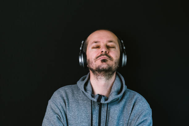 bearded relaxed man in hoodie standing against black background with headphones and listening music - head and shoulders audio imagens e fotografias de stock