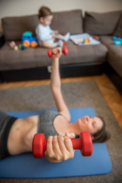 woman working out with dumbbells in her living room while toddler boy sitting on sofa and playing with toys - mother exercising baby dieting imagens e fotografias de stock