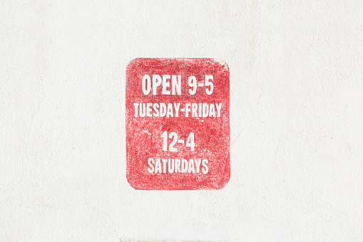 A red sign with white letters is painted on a white wall explaining the opening hours of the business.