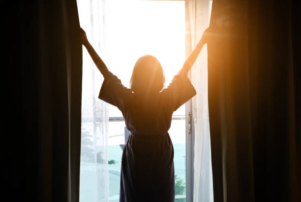 la mujer se despertó temprano. se paró en la ventana de su dormitorio y levantó ambos brazos para refrescarse. por la mañana - arms lifted fotografías e imágenes de stock