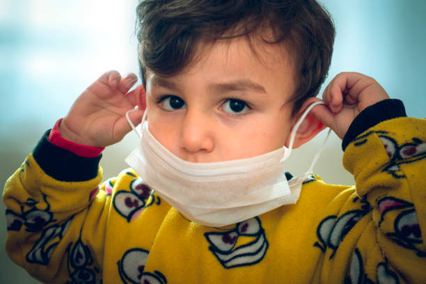 Funny looking 3 years old curious baby in pajamas trying to wear surgical mask stock photo