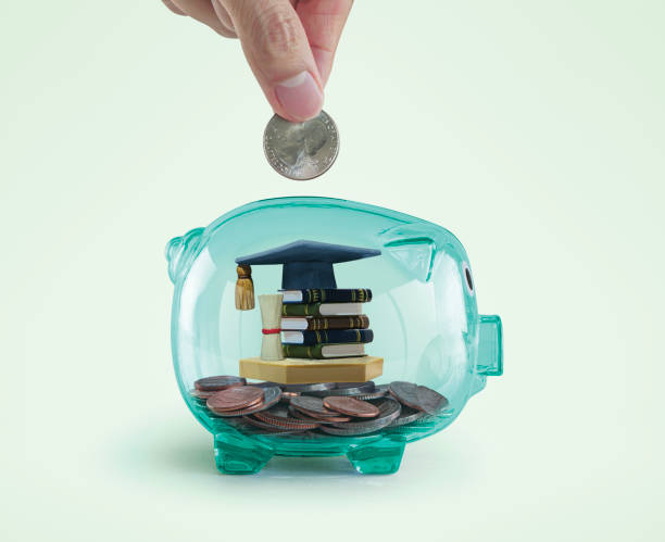 Money savings for education concept. Hand putting a coin into a green piggy bank with books, diploma and graduation cap toy inside. stock photo