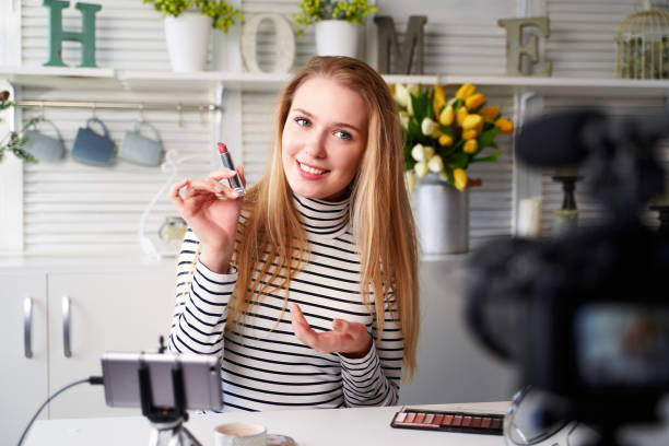 istock Vlogger female showing lipstick. Beauty blogger woman filming daily make-up routine tutorial near camera on tripod. Influencer blonde girl live streaming cosmetics product review in home studio 1218340616