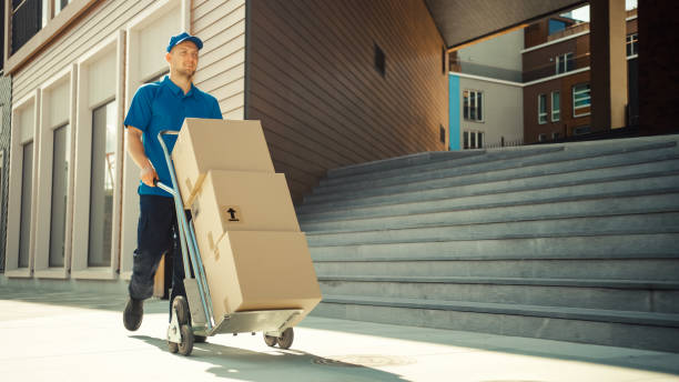 feliz joven repartidor empuja carro de camión de mano lleno de cajas de cartón y paquetes para la entrega. mensajero profesional trabajando eficiente y rápidamente. en el fondo elegante área urbana moderna - carretilla industrial fotografías e imágenes de stock