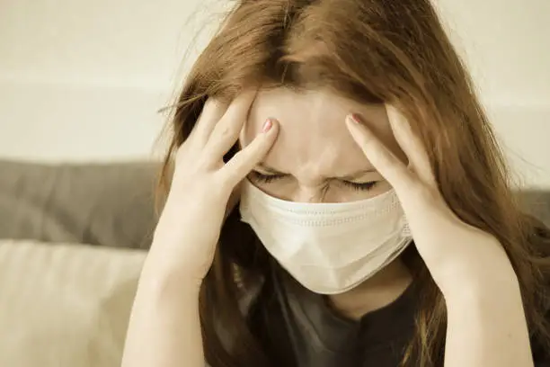 Photo of A redhead girl in a medical mask has a headache. Woman with a sore head sits on the bed