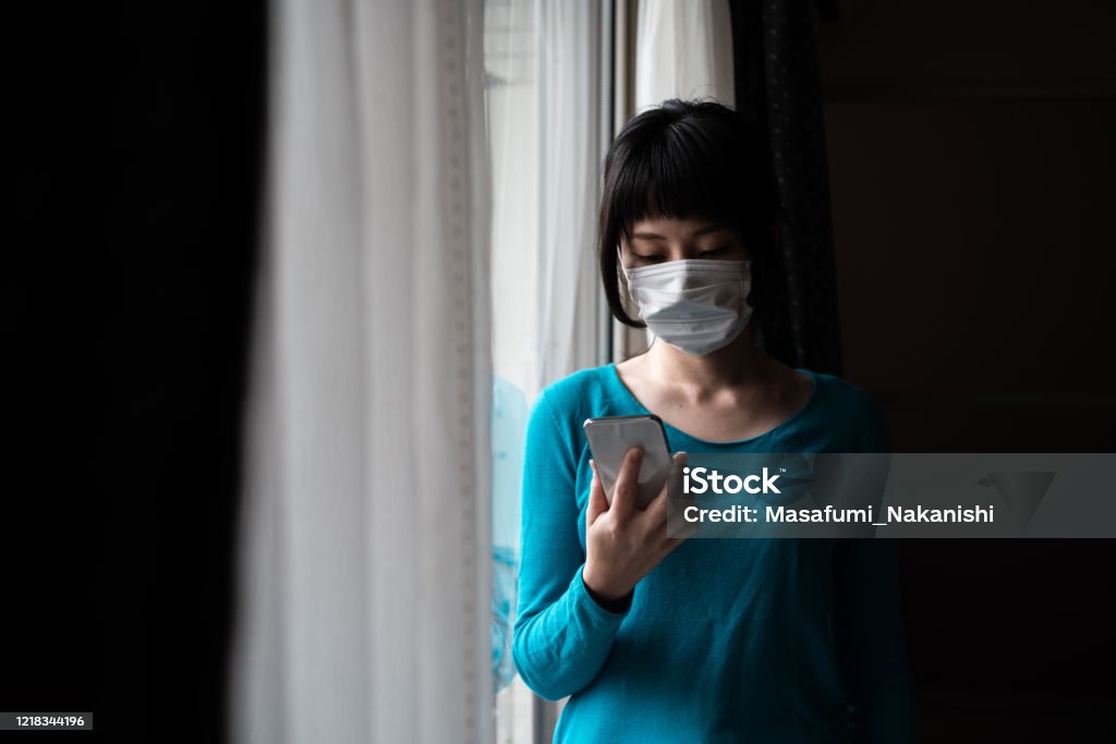 窓にスマートフォンを操作する外科マスクを身に着けているアジアの女性の屋内肖像画 - 日本人のロイヤリティフリーストックフォト