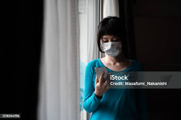 窓にスマートフォンを操作する外科マスクを身に着けているアジアの女性の屋内肖像画 - 日本人のストックフォトや画像を多数ご用意 - 日本人, コロナウイルス, 心配する