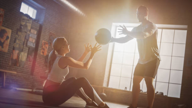 istock Young Girl Exercises with Personal Trainer, Doing Sit-ups with Medicine Ball, Throwing Pass Back and Forth. Strong Couple Workout. Exercising Strength, Cardio and Power. Shot with Warm Sun Flare. 1218344514