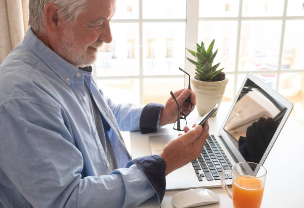 l’homme aîné barbu reste à la maison utilisant le téléphone cellulaire. bureau de bureau avec ordinateur portable et verre de jus d’orange. les aînés et le concept technologique. lumière intense de la fenêtre - telephone keypad old white photos et images de collection