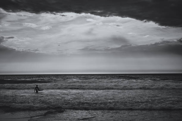 istock Lone surfer walks into the ocean to catch a wave. View on an Atlantic coast and dramatic cloudy sky at sunset. Concept of outdoor activities. Lisbon, Portugal. High Contrast. B&W color. 1218356038
