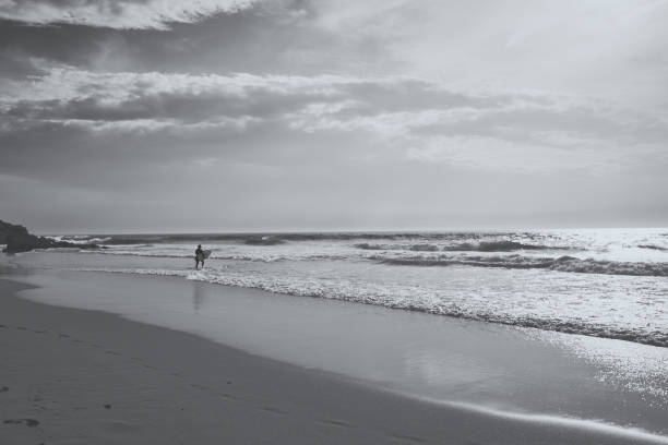 istock Lone surfer walks into the ocean to catch a wave. View on an Atlantic coast and dramatic cloudy sky at sunset. Concept of outdoor activities. Lisbon, Portugal. High Contrast. B&W color. 1218356052