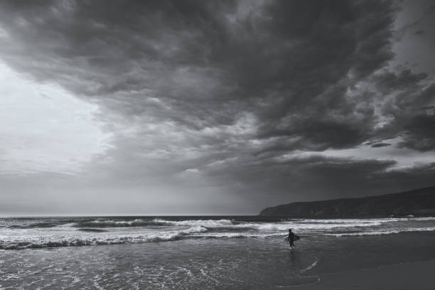 istock Lone surfer walks into the ocean to catch a wave. View on an Atlantic coast and dramatic cloudy sky at sunset. Concept of outdoor activities. Lisbon, Portugal. High Contrast. B&W color. 1218356056
