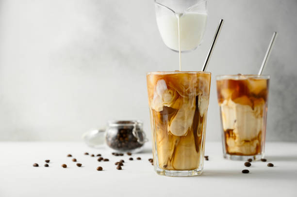 o leite é derramado no café com gelo em um fundo leve. dois copos transparentes de café gelado refrescante em uma mesa branca. - milk shake coffee latté cold - fotografias e filmes do acervo