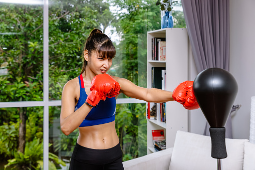 Female woman boxer hitting punching bag in living room at home, practice martial arts self-defense
