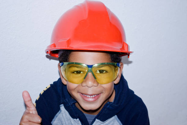 Yellow safety goggles, orange gloves, and a cordless drill rest on a wooden surface outdoors, surrounded by greenery. Tools for construction or DIY projects