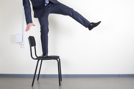 Businessman balancing while standing on an office chair against the wall with space for text.