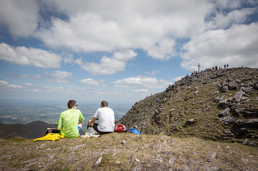 Two hikers resting and eating at Carauntoohil Mountain, County Kerry, Ireland, on a hot summer day with blue skies and patchy cloud, with the summit in the distance