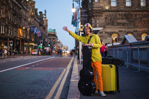 Traveling musician with a yellow suitcase and a guitar case waiting for a taxi ride on a sidewalk.