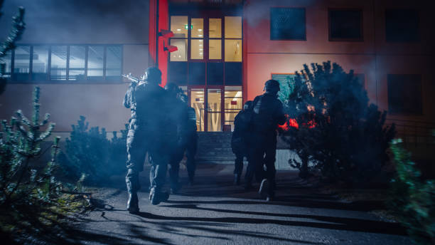maskiertes team bewaffneter swat-polizisten läuft in formation vorwärts ein bürogebäude. soldaten mit gewehren und taschenlampen laufen auf einer straße voller rauch. - spezialeinheit stock-fotos und bilder
