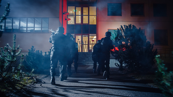 Masked Team of Armed SWAT Police Officers Run in Formation Forwards an Office Building. Soldiers with Rifles and Flashlights Run on a Street Filled with Smoke.