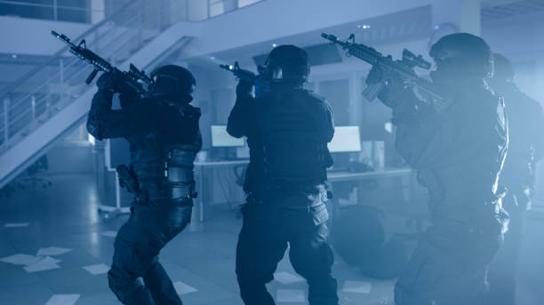 equipe mascarada de policiais armados da swat move-se lentamente em um salão de um edifício de escritórios apreendidos escuros com mesas e computadores. soldados com rifles e lanternas vigiam e cobrem o entorno. - black ops - fotografias e filmes do acervo