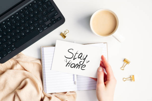 istock Flat lay feminine workspace during coronavirus COVID-19 self isolation, quarantine, lockdown. Top view laptop, coffee cup, paper notebook, blanket, female hand holding card with message 