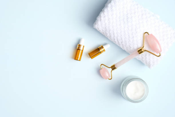 Jade roller for face massage with essential oil bottles, towel, cream moisturizer on blue background. Flat lay, top view, copy space. Rose quartz facial massage tool, beauty tool for skin care concept