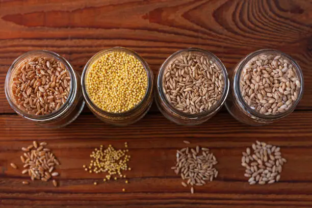 Cereal grains in glass jars on wooden background. Collection of different groats top view barley, oats, millet and wheat. Wholegrain foods with high fiber content flat lay. Healthy diet ingredients.