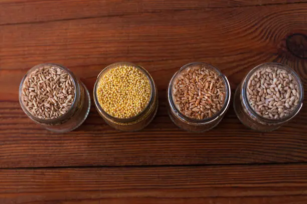 Cereal grains in glass jars on wooden background. Collection of different groats top view barley, oats, millet and wheat. Wholegrain foods with high fiber content flat lay. Healthy diet ingredients.