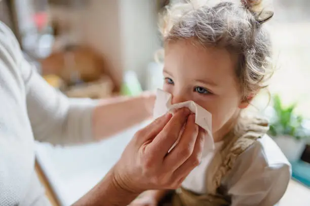 Unrecognizable father with small sick toddler daughter indoors at home, blowing nose.