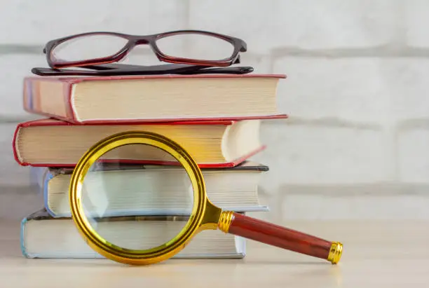 Photo of A stack of books, glasses, and a magnifying glass on the table.The concept of information search.