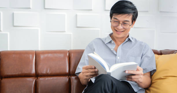 自宅のリビングルームでソファの本を読むアジアの上級男性、アジアの高齢者の肖像画は自宅で雑誌を読んでリラックスして幸せです - senior adult relaxation sofa reading ストックフォトと画像